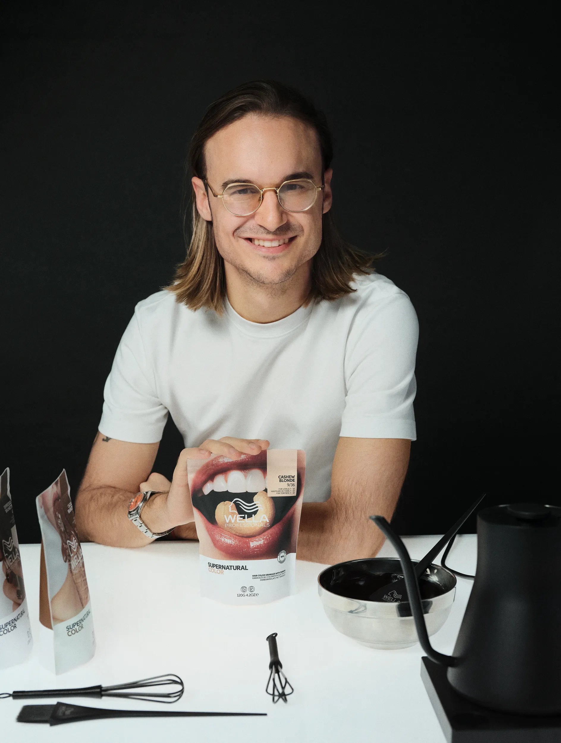 Εικόνα του Wella Professionals ambassador, Marco Firriolo, να χαμογελάει και να φοράει γυαλιά και ένα λευκό t-shirt. Ο Marco κάθεται σε ένα τραπέζι με το χέρι του σε μία συσκευασία Supernatural Color.  Πάνω στο τραπέζι υπάρχουν κι άλλες αποχρώσεις Supernatural Color και εργαλεία styling. 