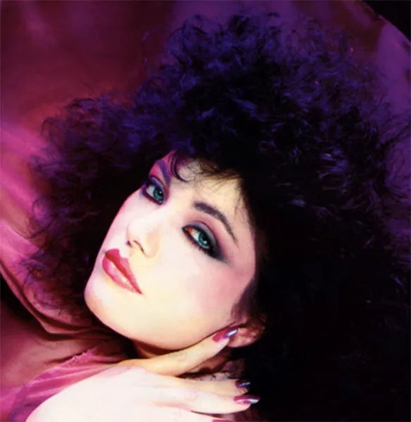 Model with volumous curly hair wearing bold make up with purple hues and, with a hand grazing her face, set against a purple background.