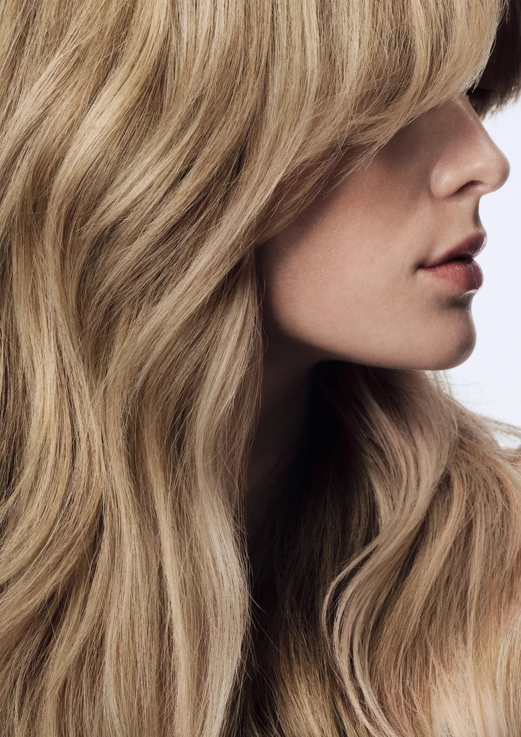 Profile view of a woman with a wavy, blonde bob hairstyle, wearing a sheer white collared shirt, against a white background.