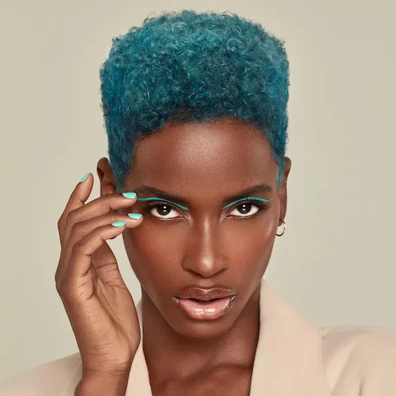 Model with short, coily, teal hair hair, green eyeshadow and green nail polish faces the camera.