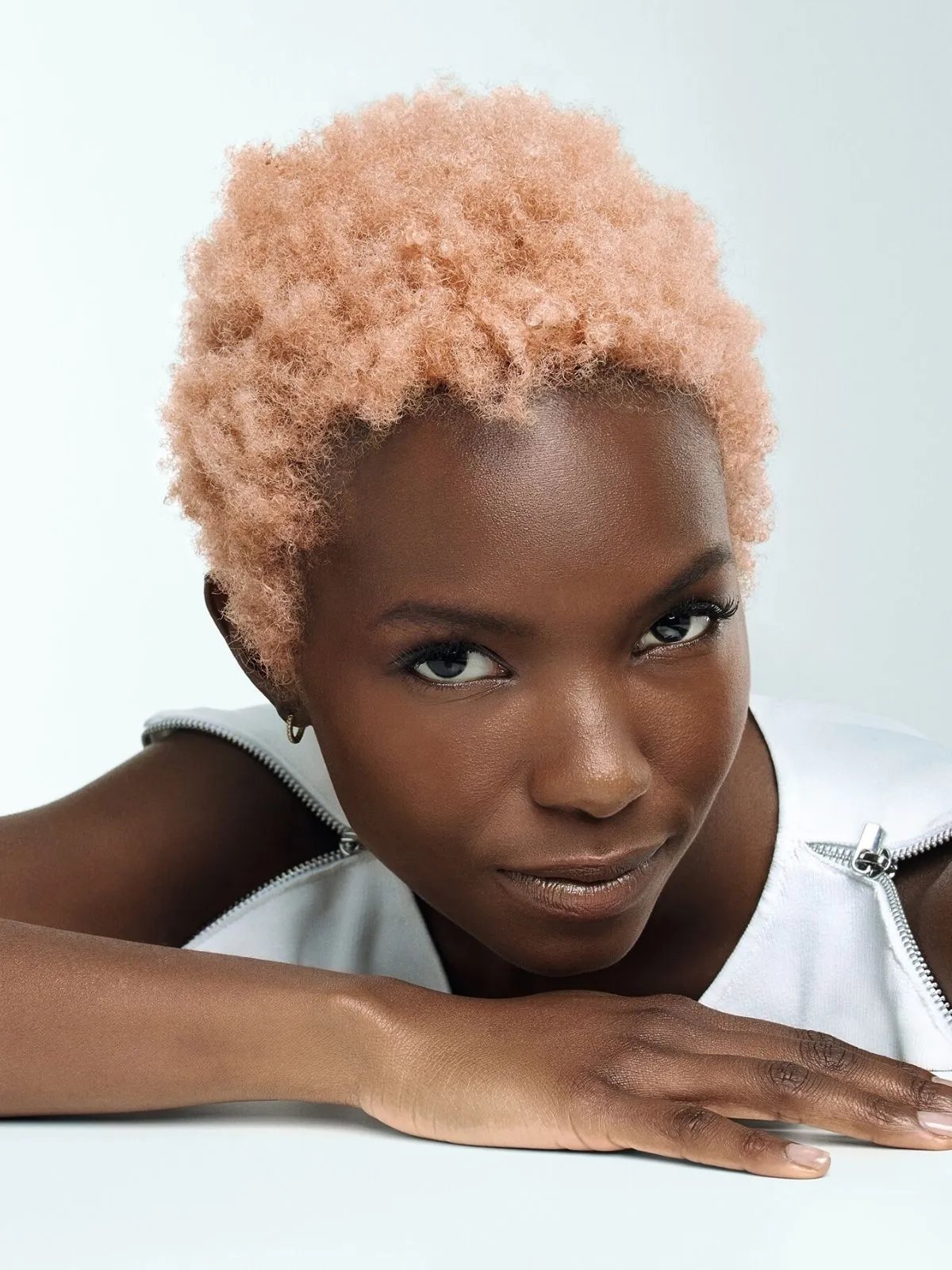 Model with rose blonde, coily hair leans against a white table.