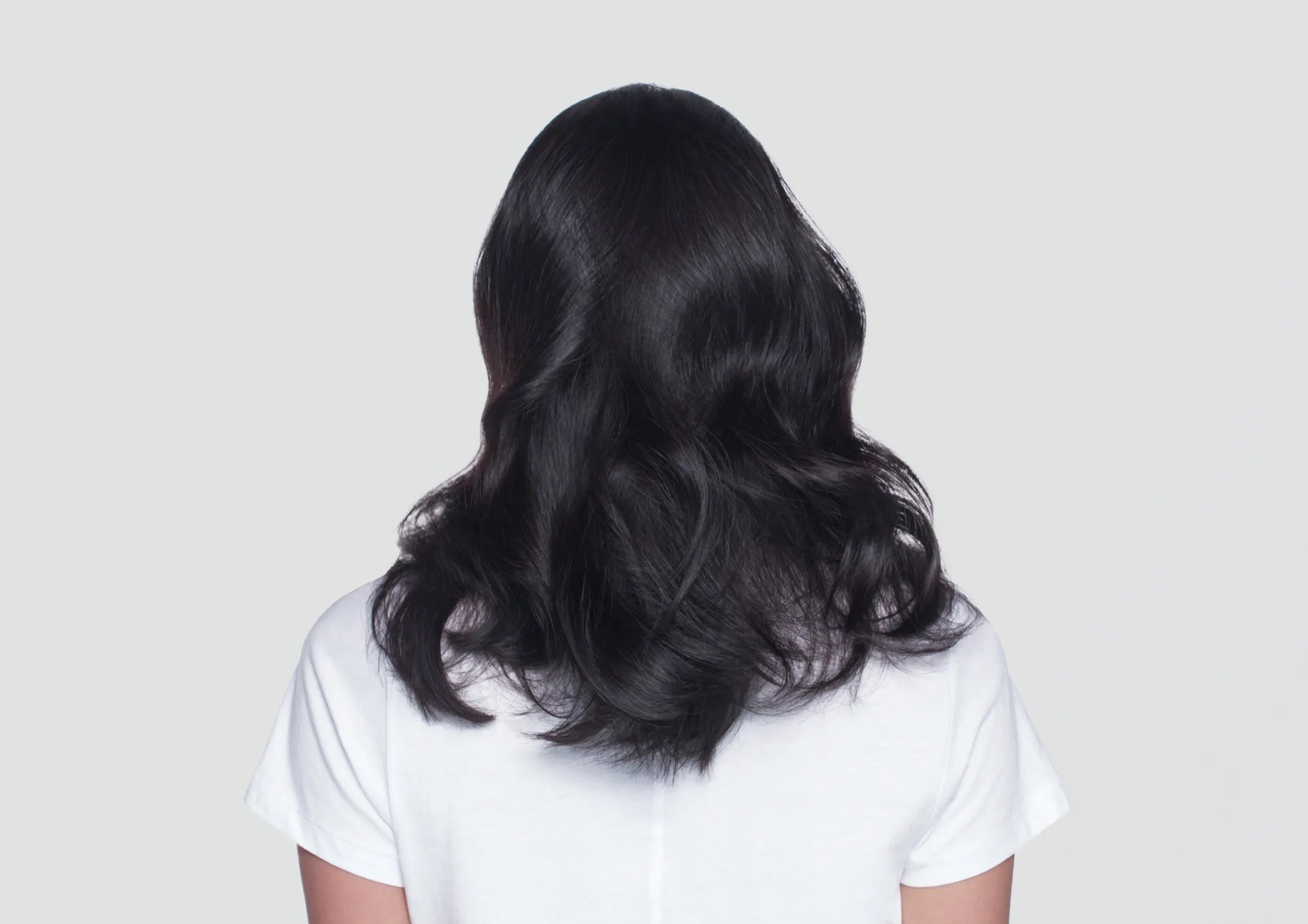 Back view of woman with shiny, wavy black hair wearing a white t-shirt on a light gray background.