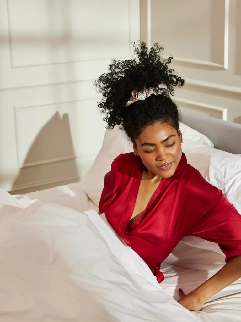 Model with dark brown, curly hair sits up in bed.