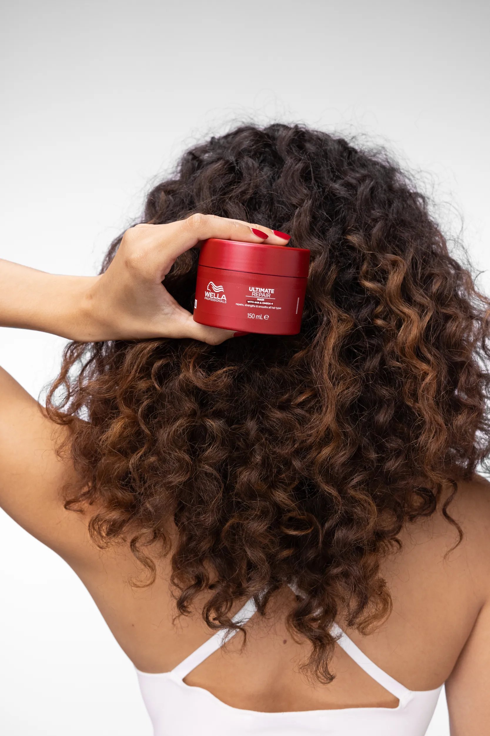Back of woman's head with brown curly hair holding ULTIMATE REPAIR Mask against a white background. 