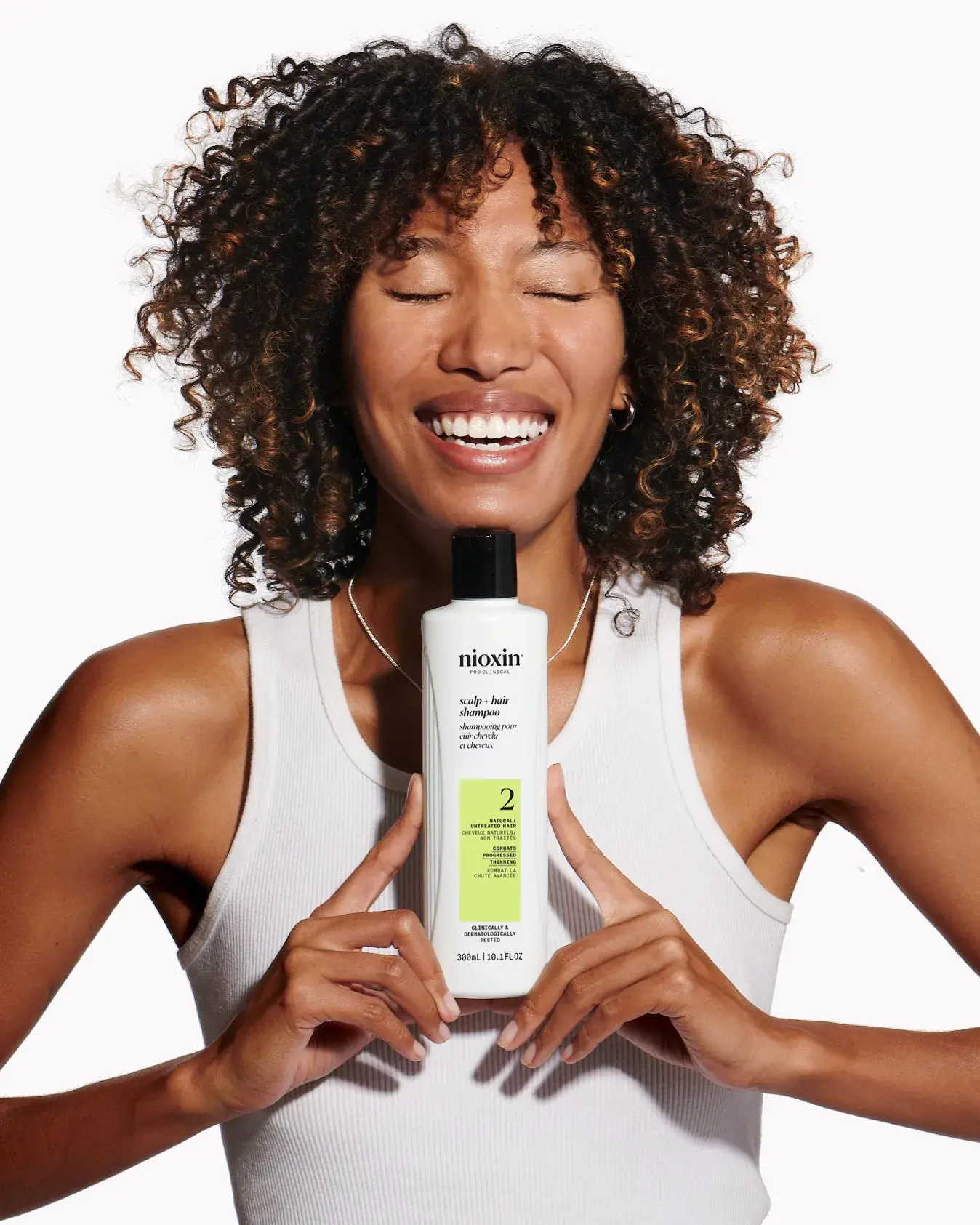 Mujer negra con el pelo rizado sonriendo y sosteniendo entre sus manos un champú para el cuero cabelludo + cabello del Sistema 4 de Nioxin
