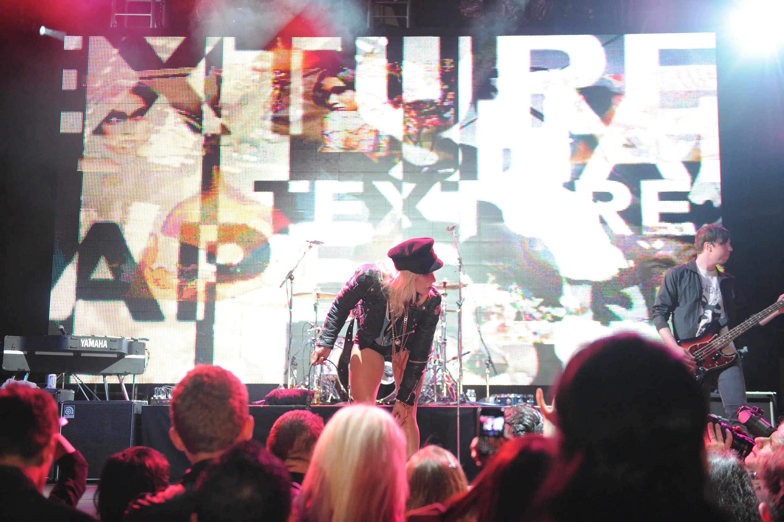 Image of woman wearing shorts, a denim jacket and a black hat, performing to a crowd. A man playing the guitar is beside her. 