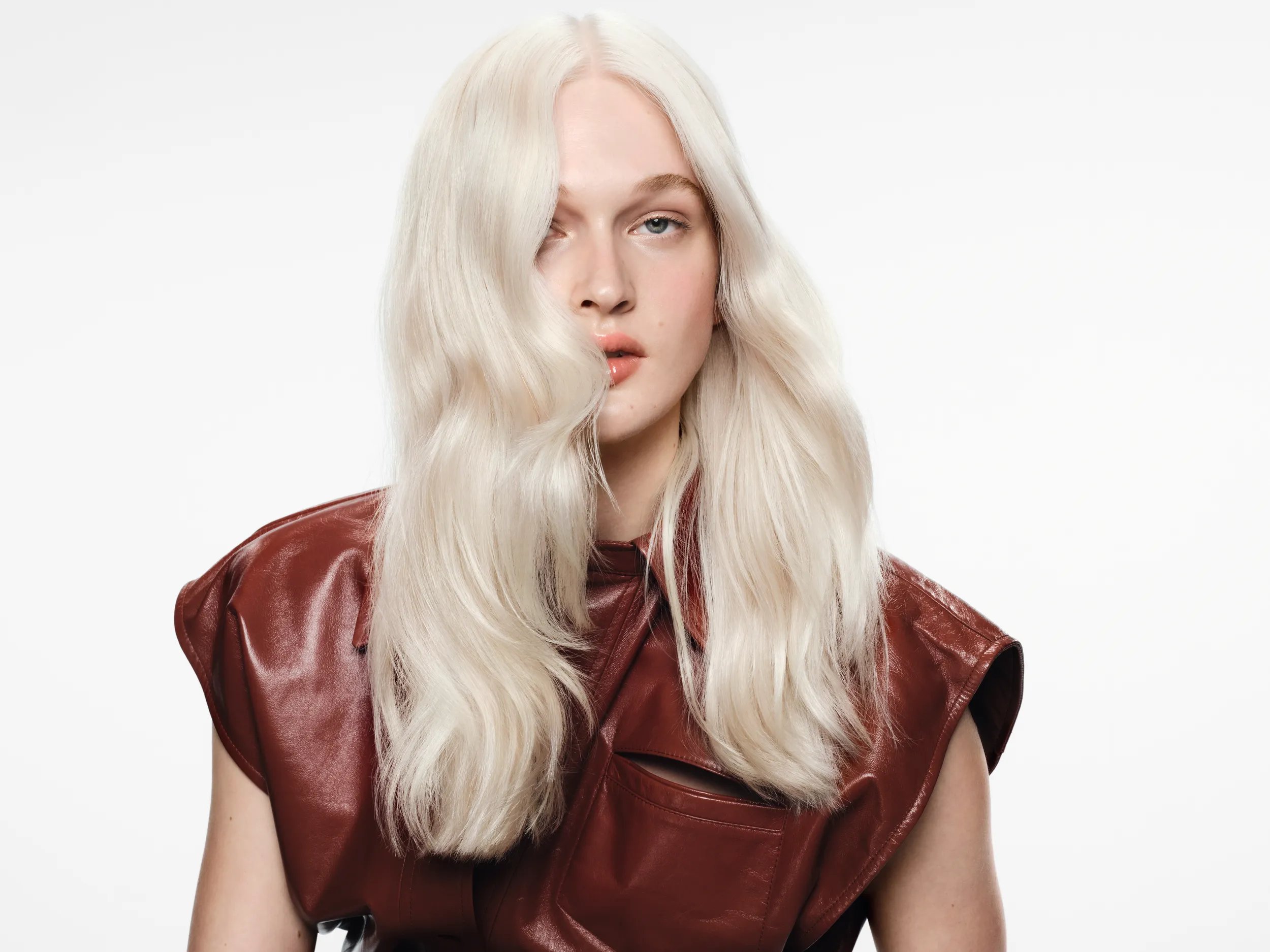 Model with long, wavy platinum blonde hair wearing a sleeveless brown leather top, looking at the camera against a plain background.