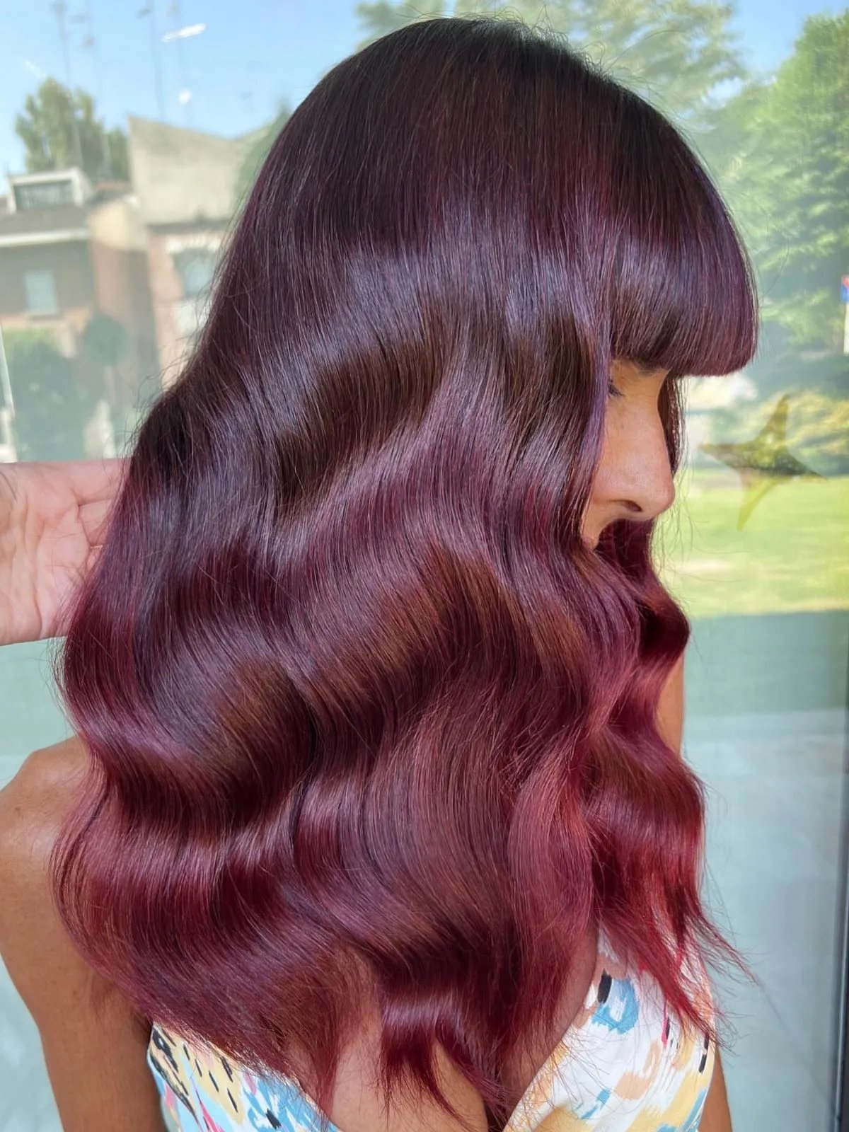 Side profile showcasing long, glossy waves in a deep burgundy-red hair color with subtle violet undertones. The hair features a full fringe and soft, defined S-shaped waves that create dimension and shine. Photographed in natural daylight against a blurred outdoor background, highlighting the rich color blend and smooth finish.