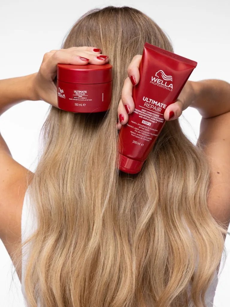 Model holds ULTIMATE REPAIR Mask and ULTIMATE REPAIR Conditioner in front of their blonde hair.