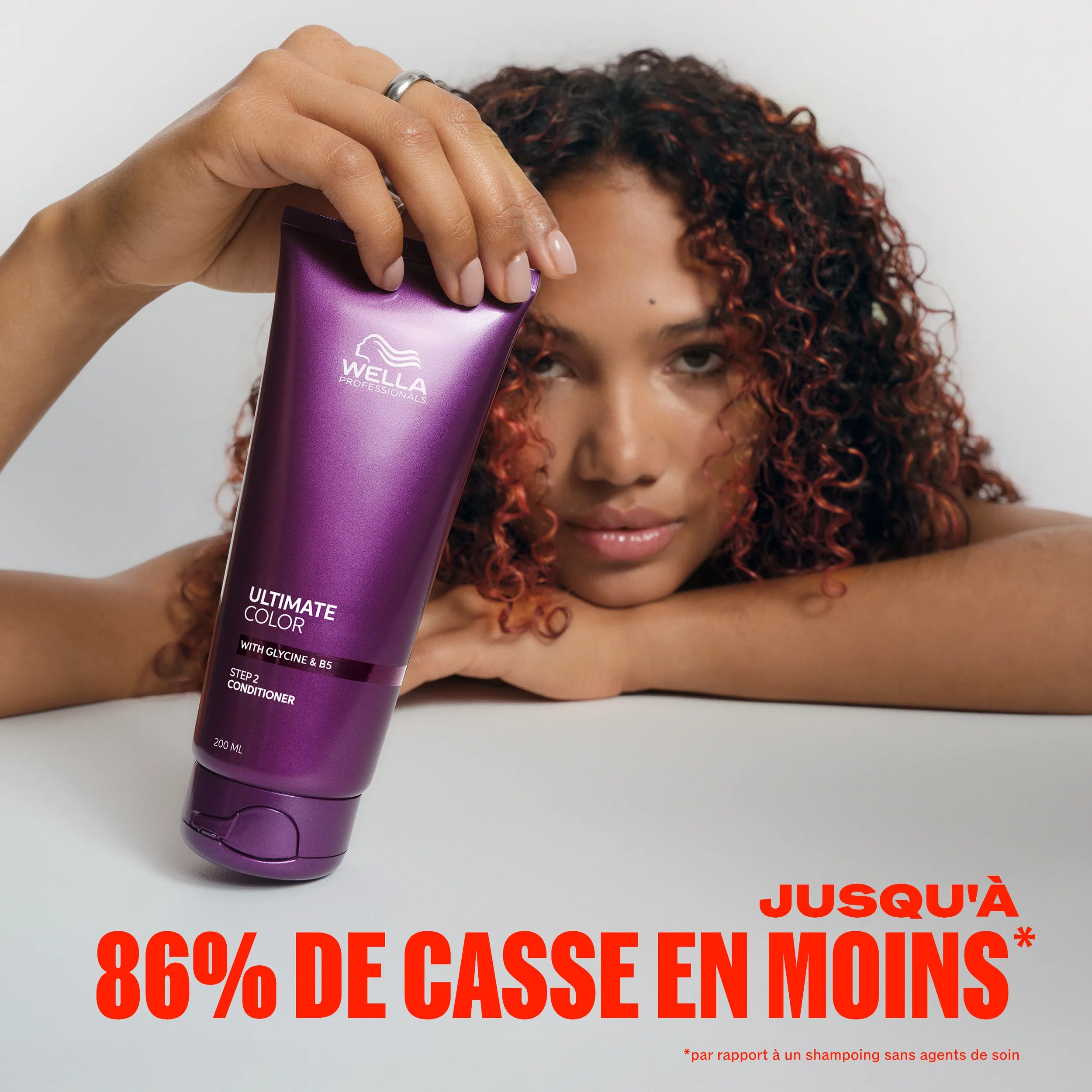 Image d’un modèle aux cheveux bruns bouclés avec des mèches rouges, appuyé sur le menton avec une main et tenant le conditioner Ultimate Color de l’autre.
Une incrustation de texte indique : "Jusqu'à 86% de casse en moins** par rapport à un shampoing sans agents de soin"		
