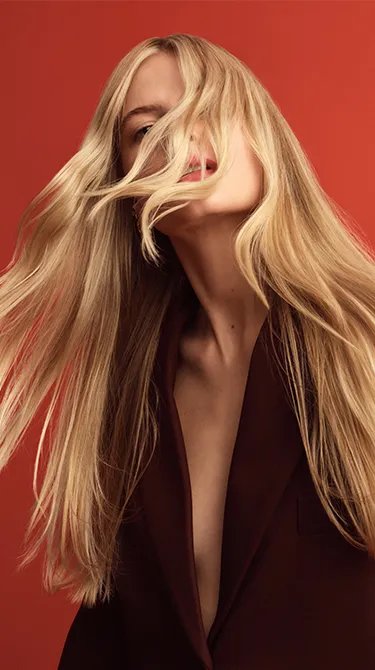 Portrait of a woman with long blonde hair partially obscuring her face, against a red background.
