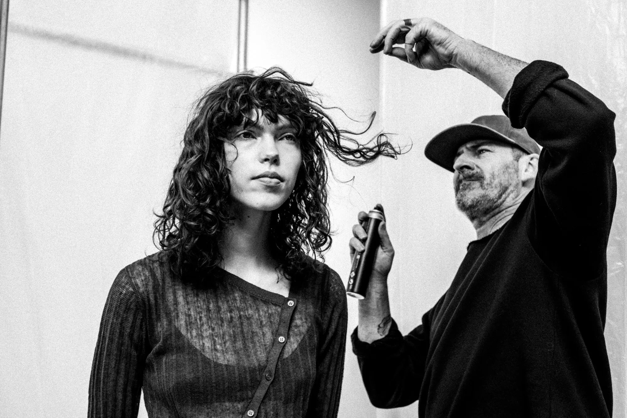 Black and white image of a hairstylist using hairspray to style a model’s curly hair during a photoshoot