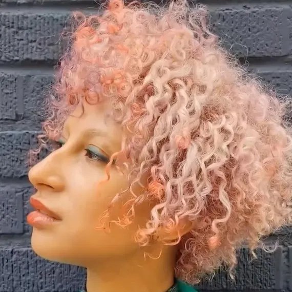 Model with short, curly, pastel pink and peach hair looks off to the side.