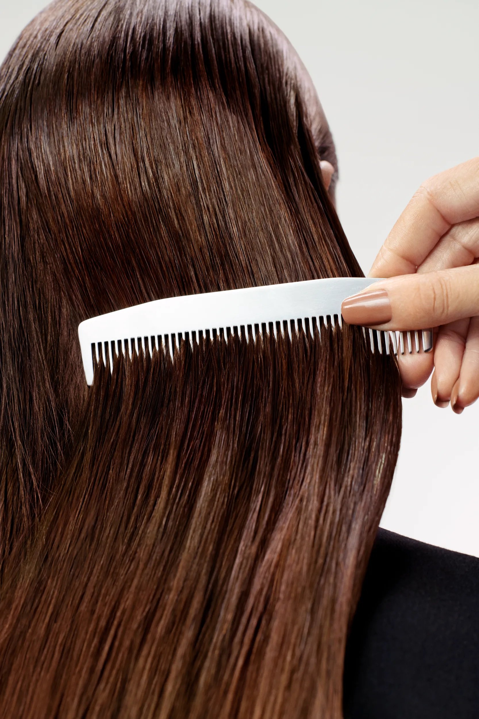 Image showing a model facing away with only her brunette hair in view. A hand is combing through her hair using a white comb. 