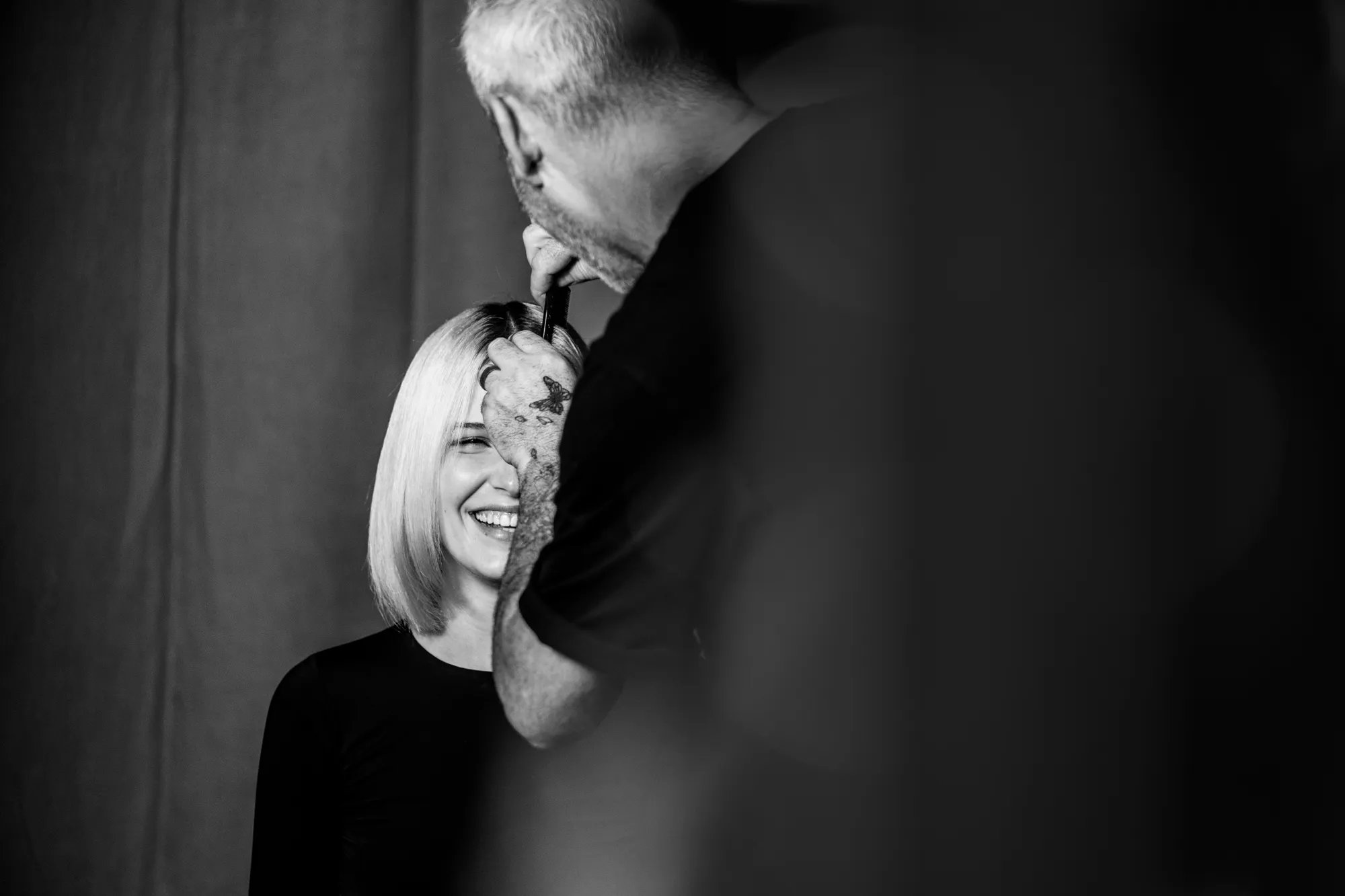 Black and white image of a model and hairstylist. Stylist is stood and combing the model's hair while the model is sat down and smiling