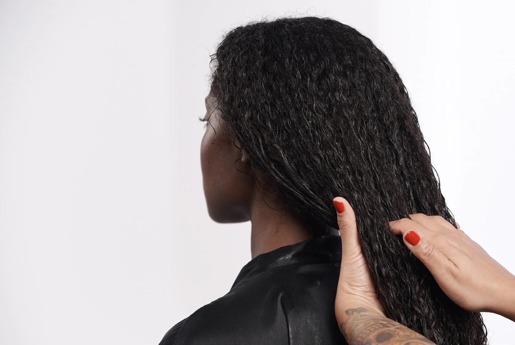 A close-up of a person with wet, curly black hair being gently styled by another person with red-painted nails and a tattooed arm.