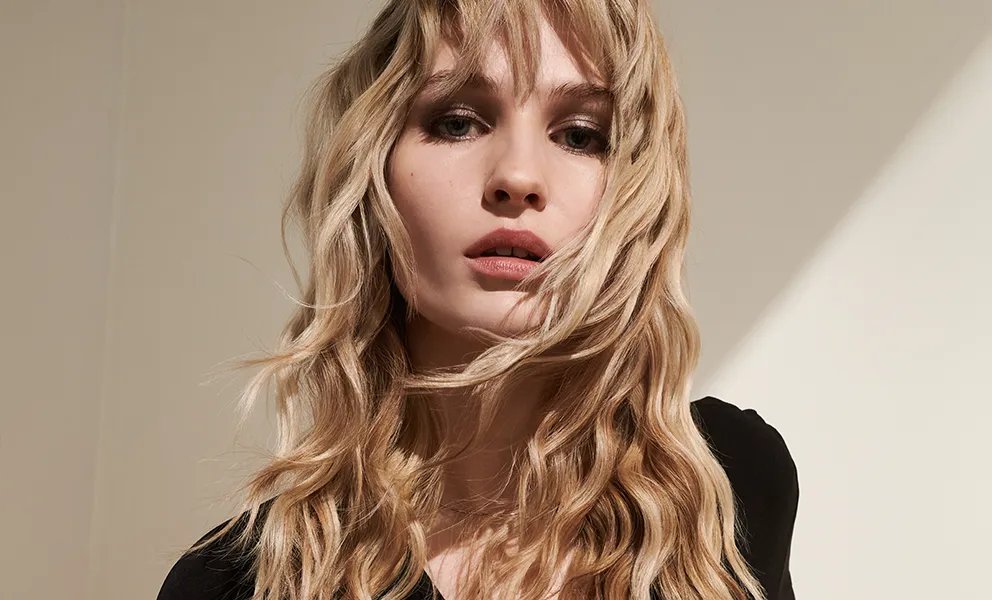 Model with sandy blonde, wavy hair and a fringe faces the camera.