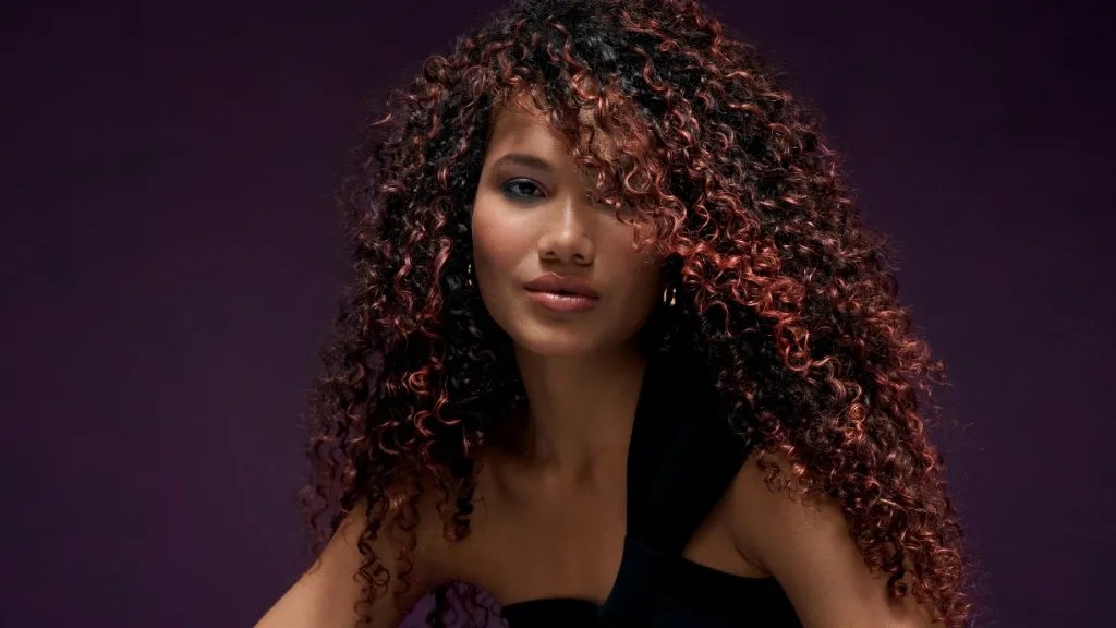 Model with long, dark brown, curly hair and red highlights.