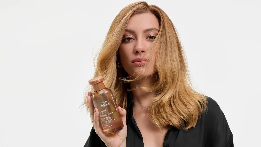 Model with silky blonde hair holds up a bottle of ULTIMATE SMOOTH Shampoo.