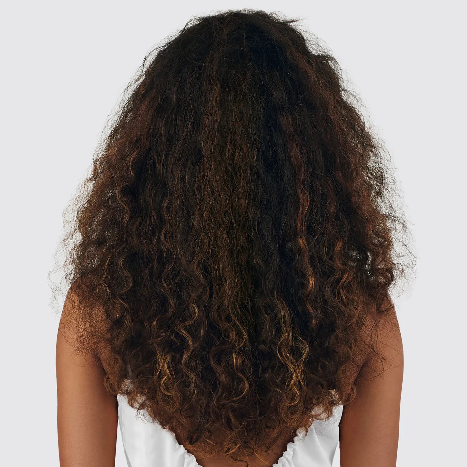 Image showing the back of a woman with long, dark, curly hair with subtle blonde highlights, wearing a white top against a neutral background.