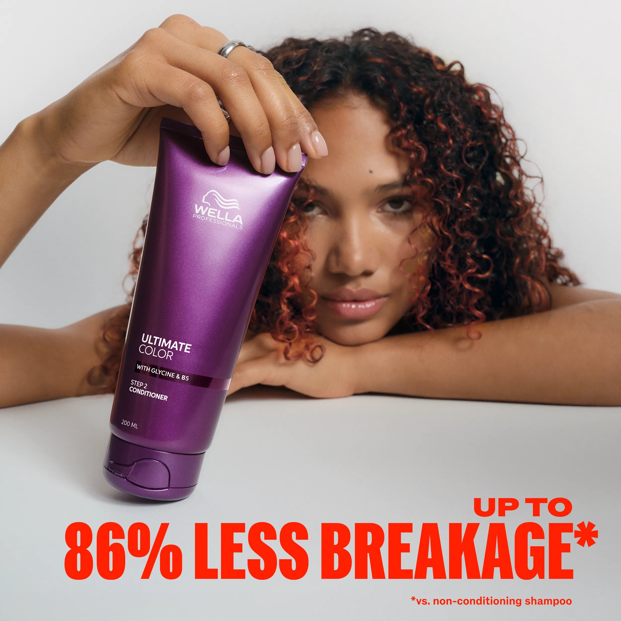 Image of model with curly brunette hair and red streaks, leaning on their chin with one hand and holding the Ultimate Color conditioner with their other. Text overlay reads "Up to 86% less breakage* *vs non-conditioning shampoo". 			

