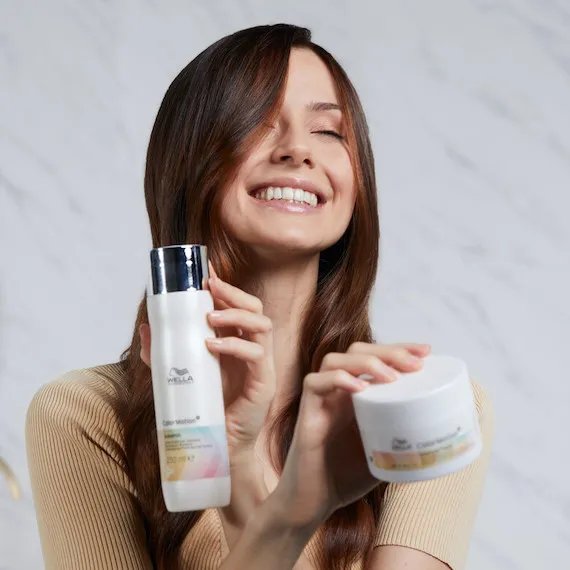 A model with long, brown hair holds a bottle of ColorMotion+ Color Protection Shampoo and a jar of the ColorMotion+ Structure+ Mask
