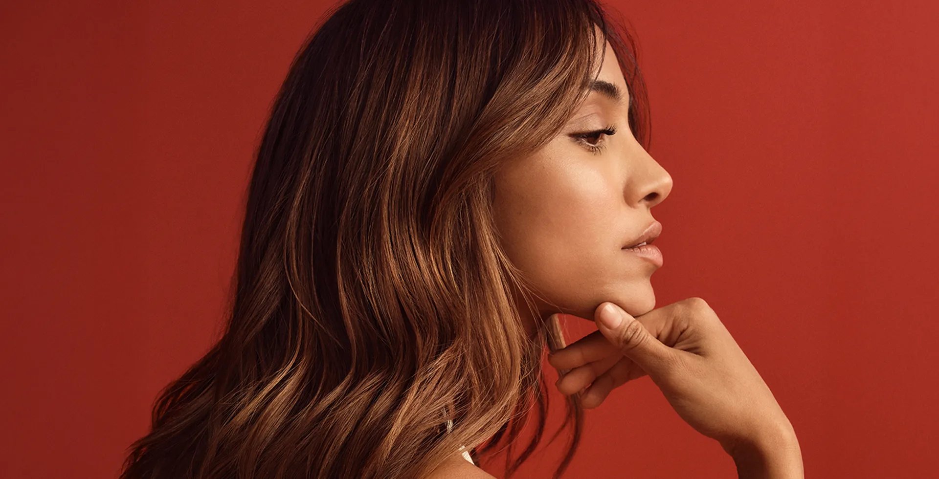 Profile portrait of a woman with wavy brown hair, resting her chin on her hand against a red background.