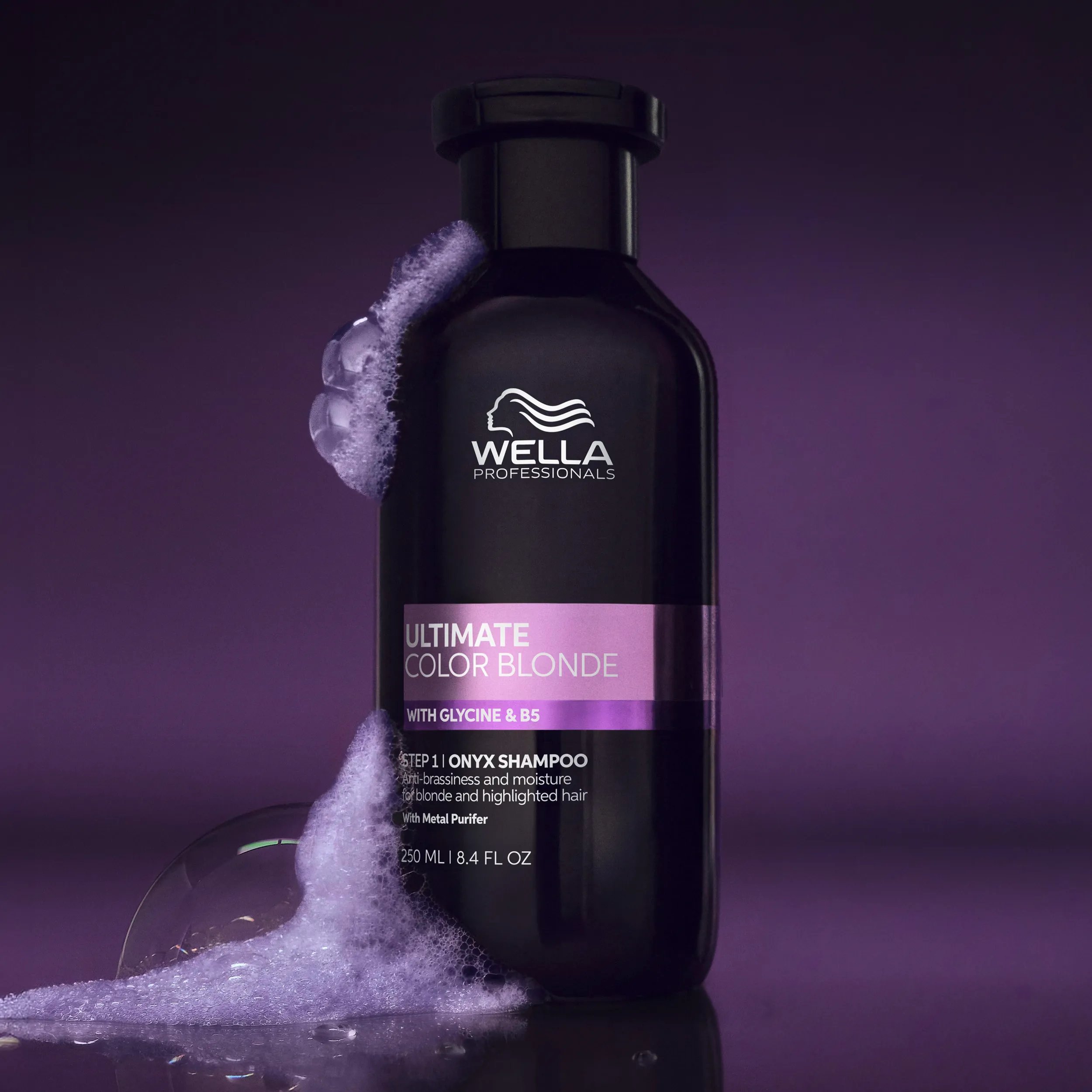 Product image of ULTIMATE COLOR Blonde Onyx Shampoo against deep purple background. Soapy bubbles cover the left side of the bottle. 