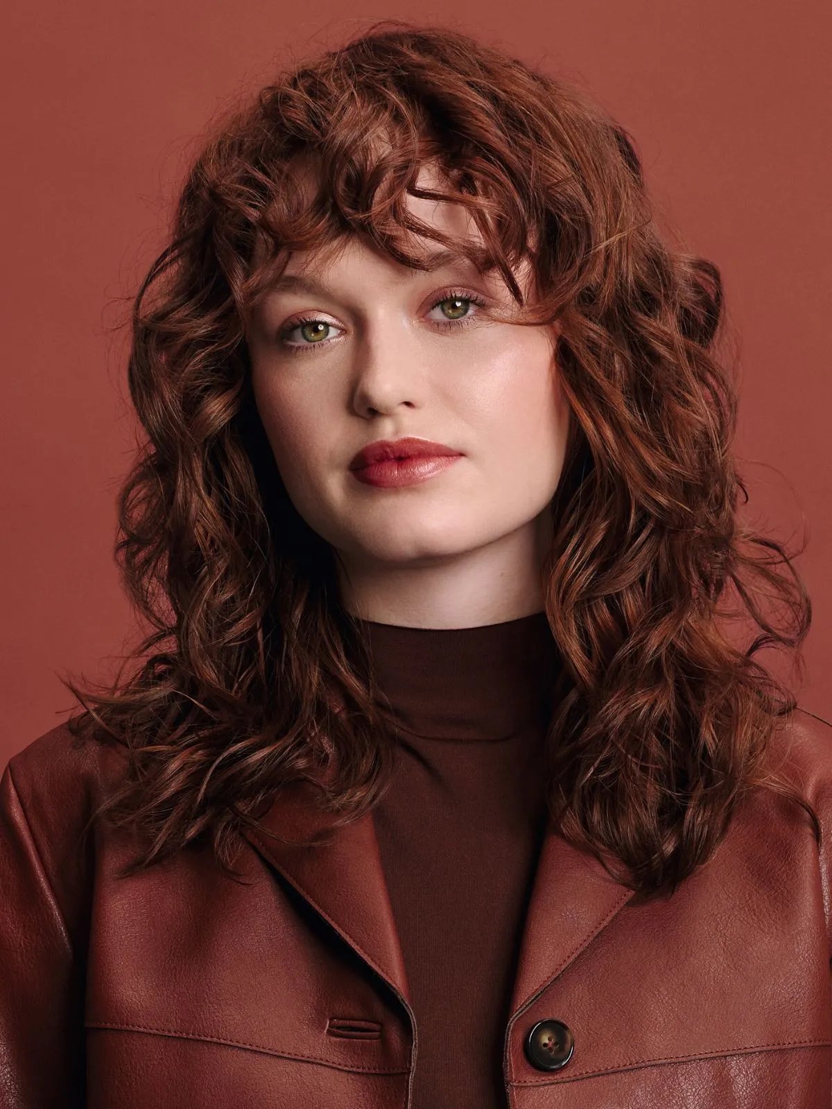 Model with red curly hair, wearing a red leather jacket against a red background. 