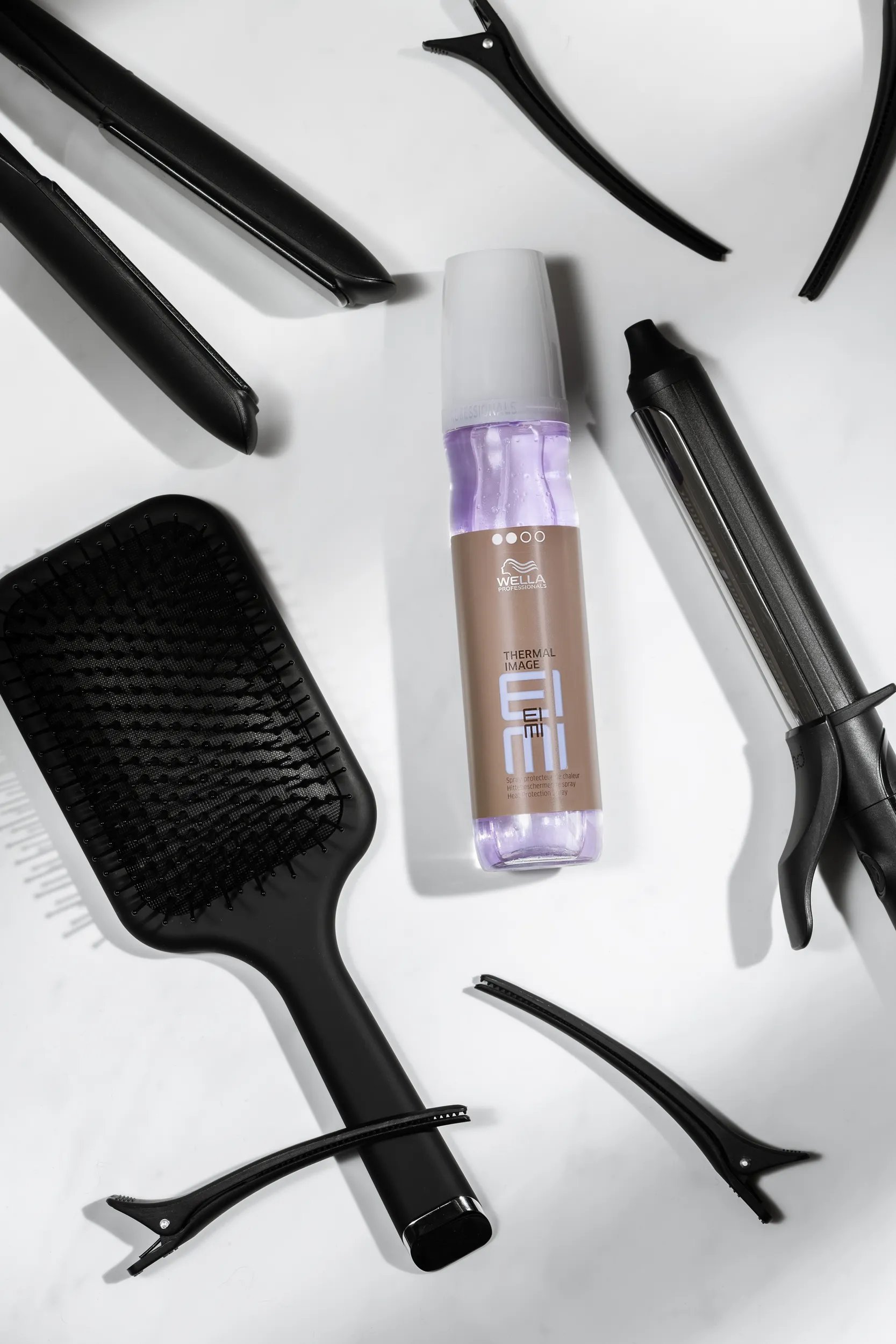 Bottle of EIMI Thermal Image heat protection spray by Wella Professionals surrounded by hair styling tools, including a brush
