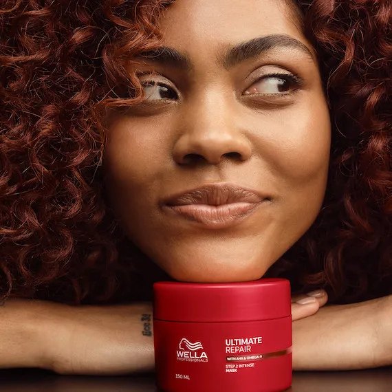 Model with dark brown, curly hair rests chin on a tub of ULTIMATE REPAIR Mask.