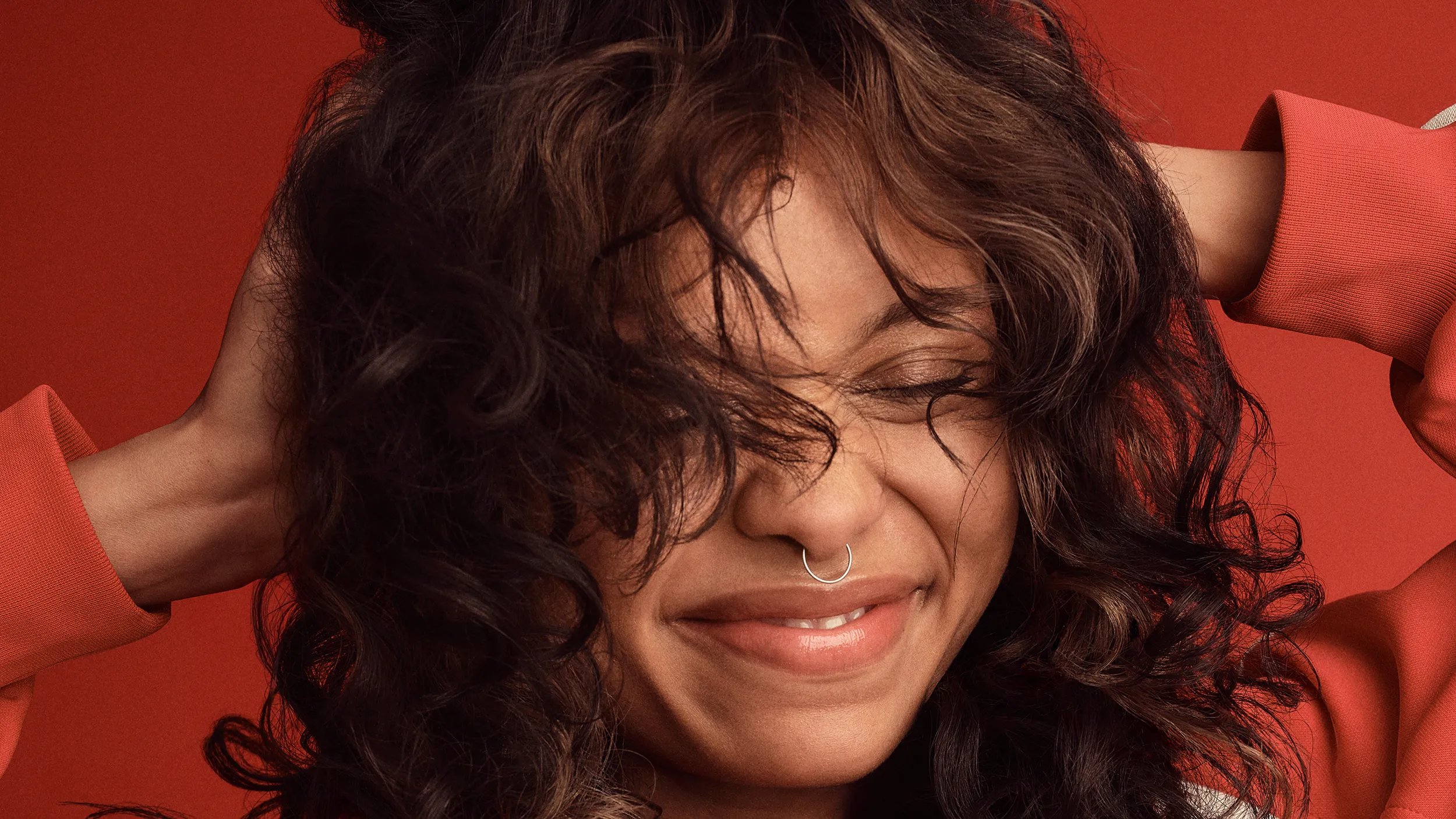 Image of a woman with curly hair, eyes closed and smiling, hands in her hair, with a vibrant red backdrop.
