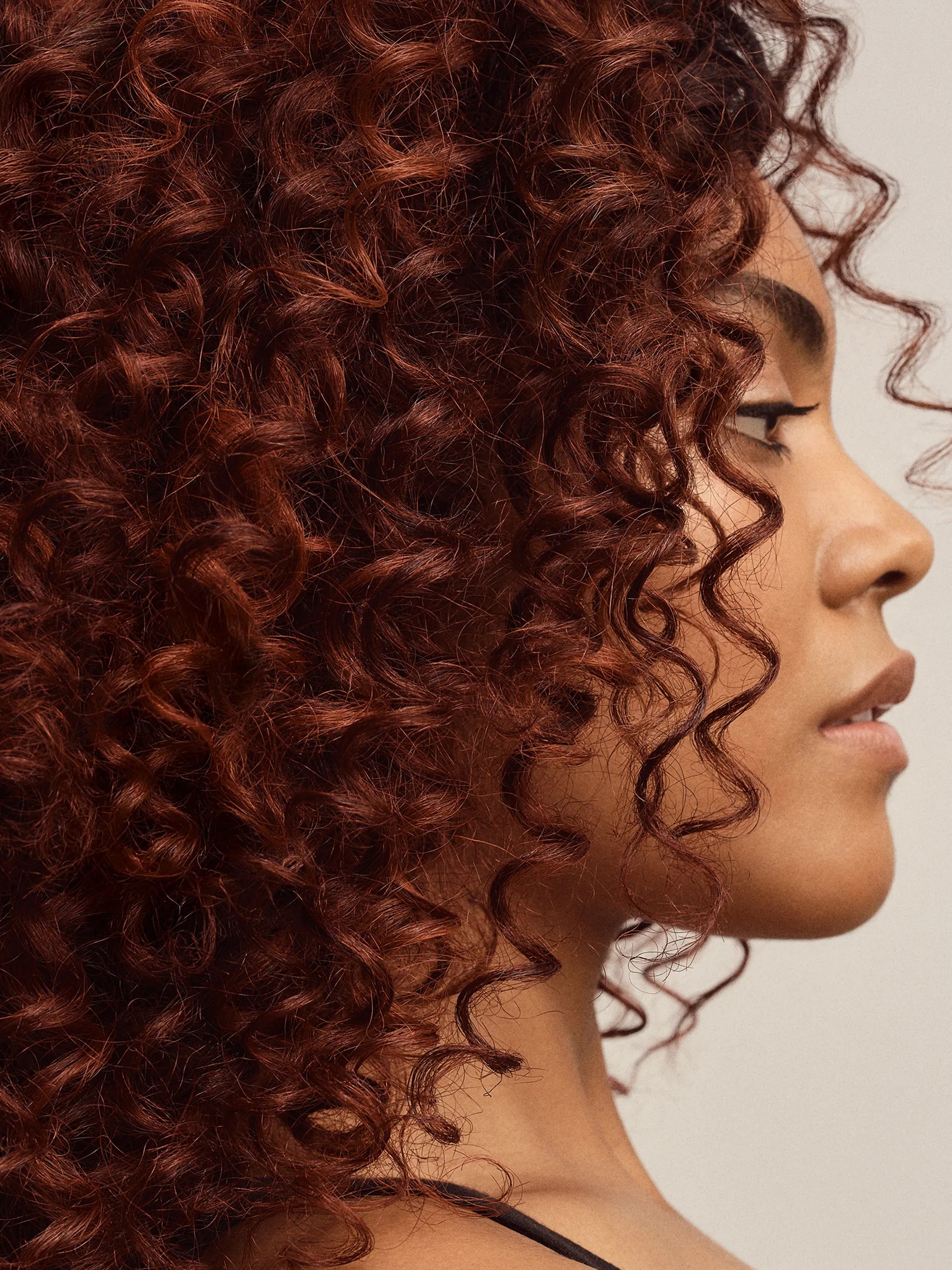 A profile view of a woman with tight, curly auburn hair against a neutral background.