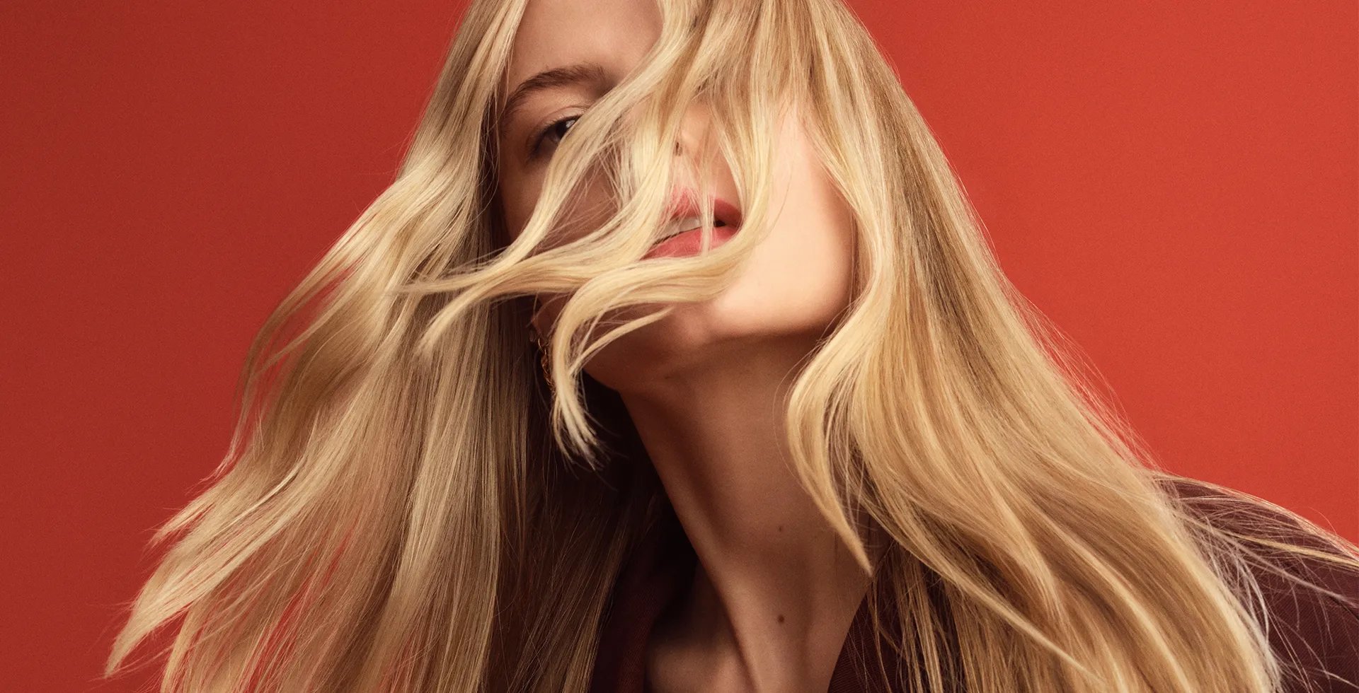 Portrait of a woman with long blonde hair partially obscuring her face, against a red background.
