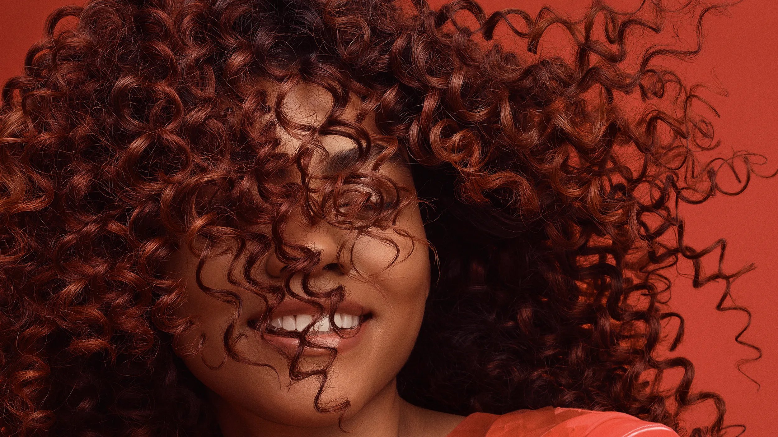 Close-up of a woman's face partially obscured by her curly auburn hair, with a bright smile against a red background.