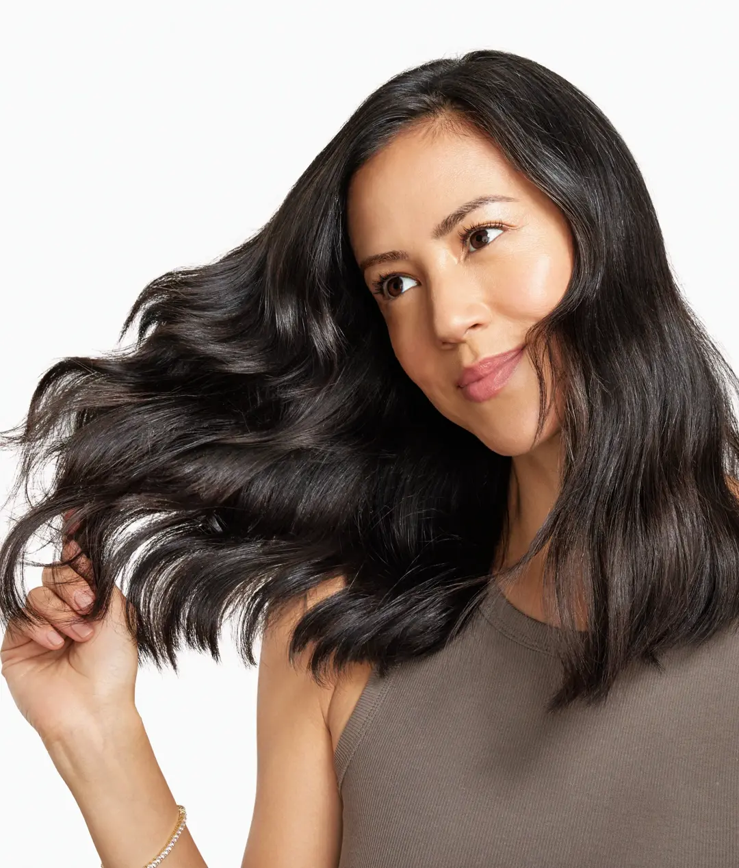 A woman in a putty-colored tank top tilts her head, smiling as she touches the ends of her mid-length, wavy hair. 