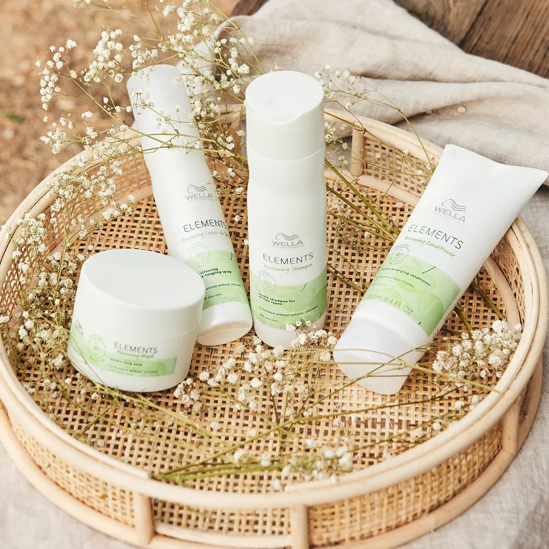 A collection of Wella Elements hair care products arranged on a woven rattan tray, surrounded by delicate white flowers, with a soft fabric and wooden surface in the background.