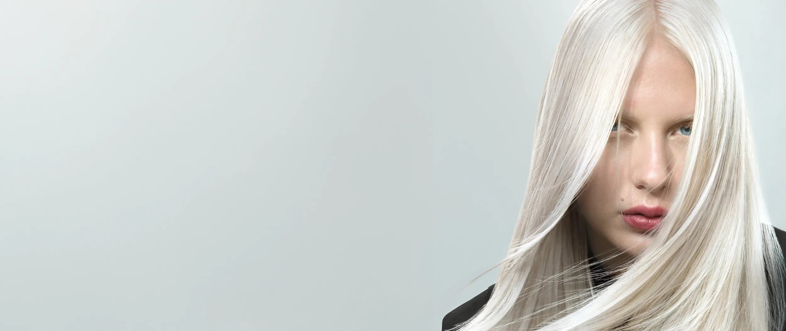 Portrait of a woman with long blonde hair partially obscuring her face, against a grey background.