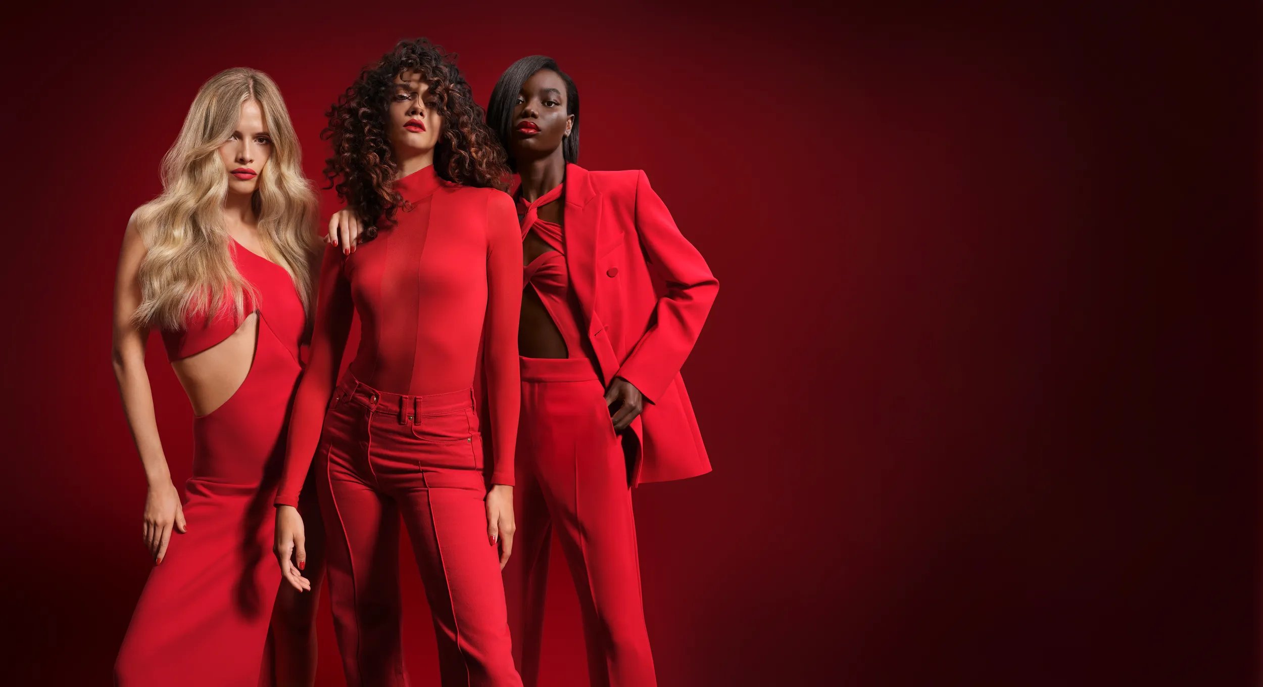 Three models pose together against a red background, dressed in red outfits. The woman on the left has long, wavy blonde hair and wears a cut-out dress. The center woman has voluminous curly hair. The woman on the right has sleek, straight black hair.