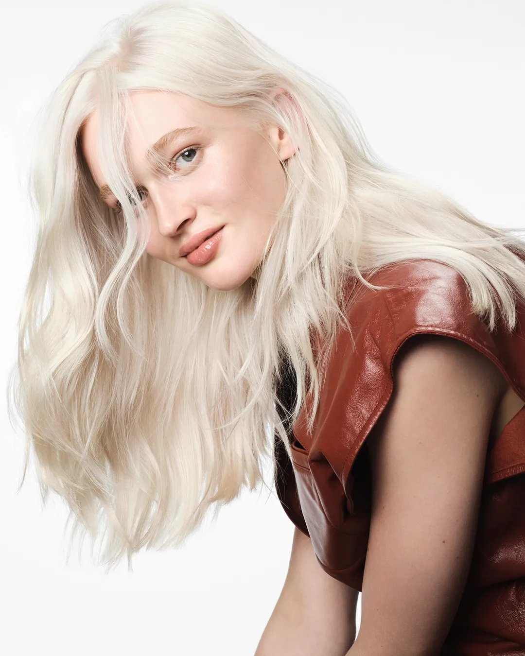 Side profile of model with white-blond and wavy hair, wearing a red sleeveless top and she is slightly smiling.