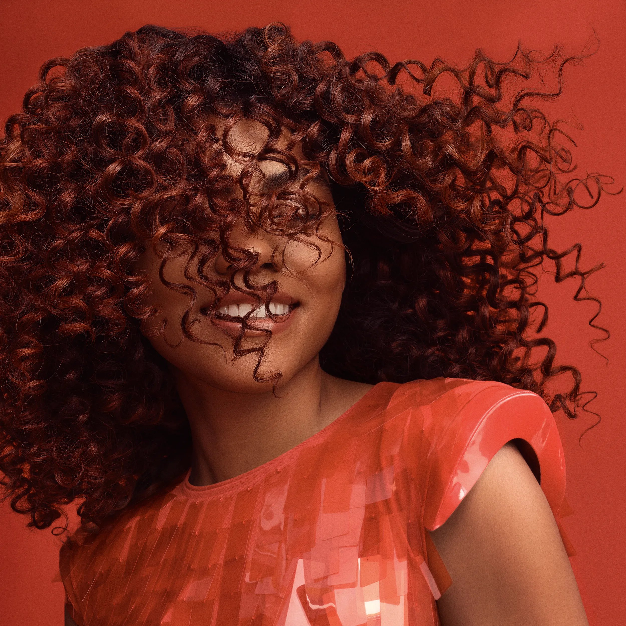 Gros plan d'une femme souriante dont les cheveux longs, bouclés et auburn cachent partiellement le visage, sur fond rouge.