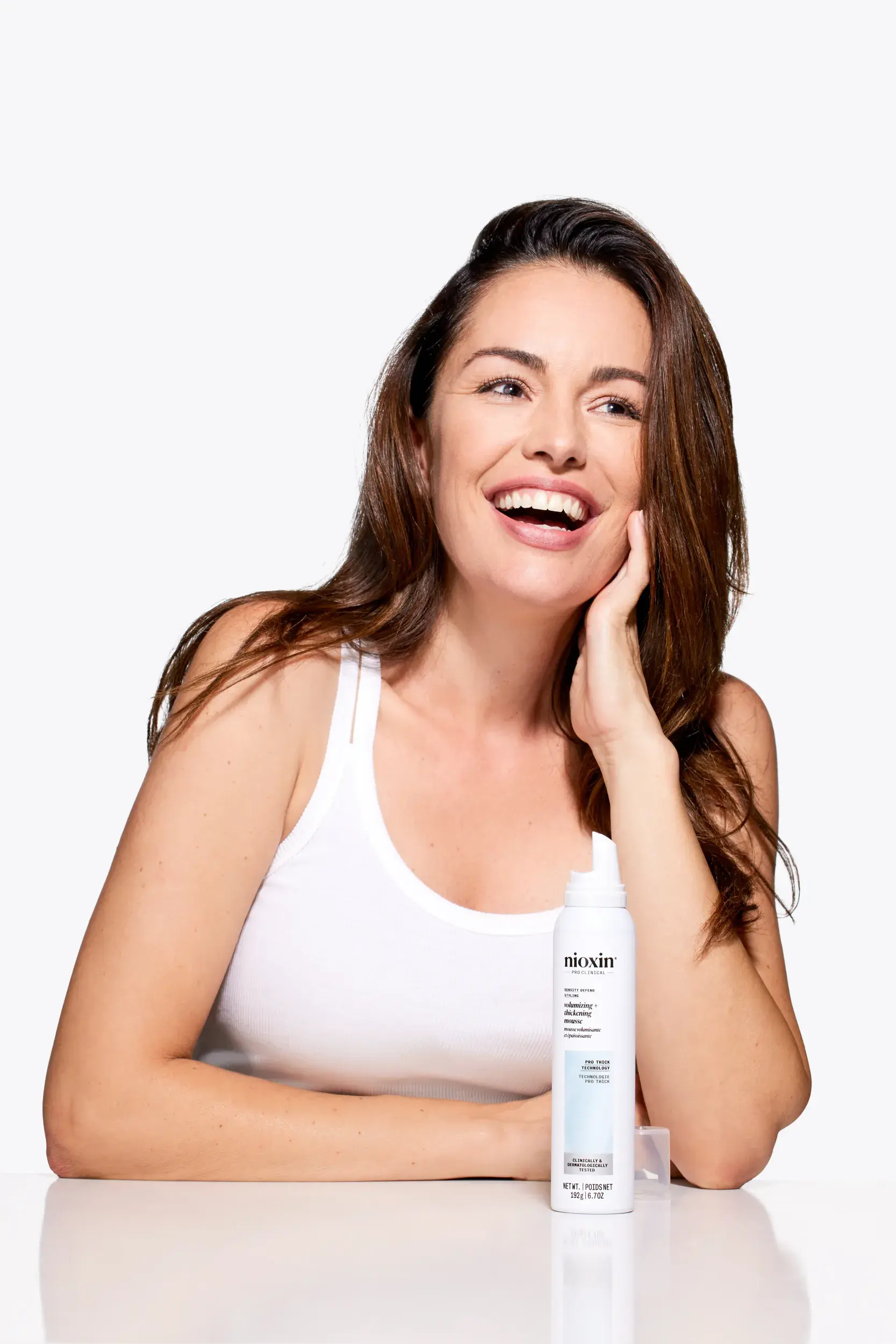 mujer morena sentada sonriendo con Nioxin Thickening Mousse en la mesa sobre fondo blanco