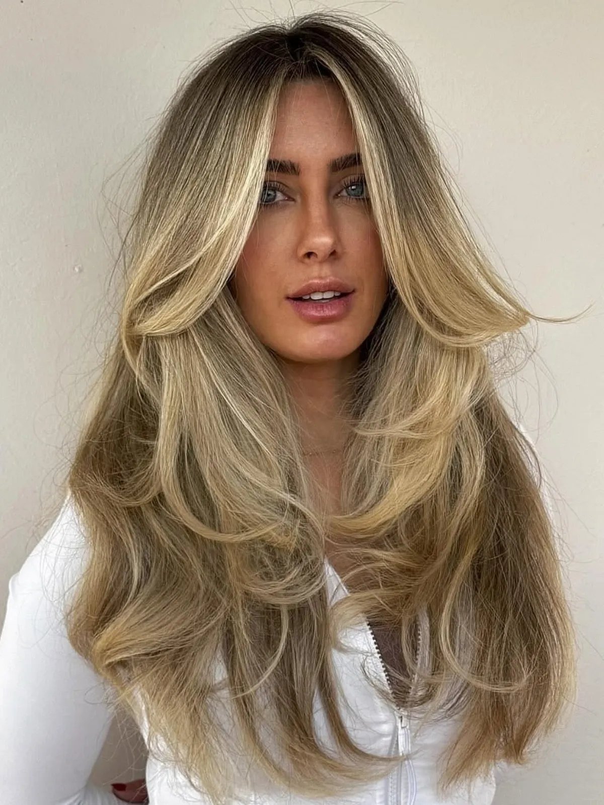Model with long, blow-dried hair featuring dark roots and neutral blonde lengths.
