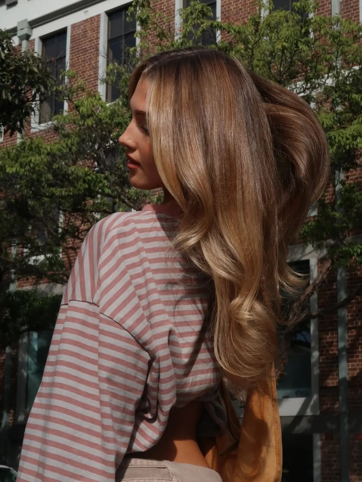 Model with long, loosely curled, light brown hair and subtle blonde accents through the ends.