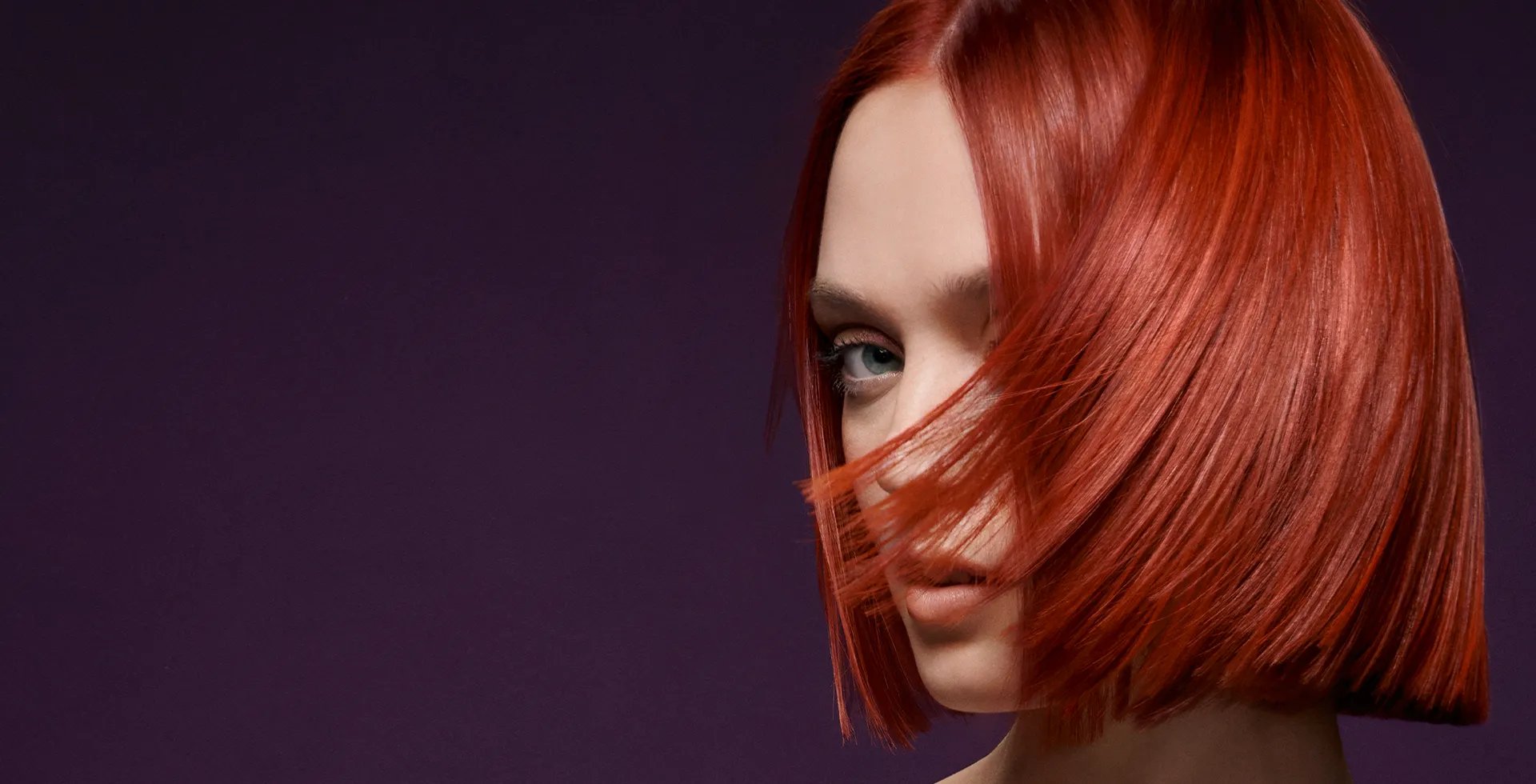 Image of model with a bright red, straight and short hairstyle against a dark purple background. Hair is slightly covering their face. 
