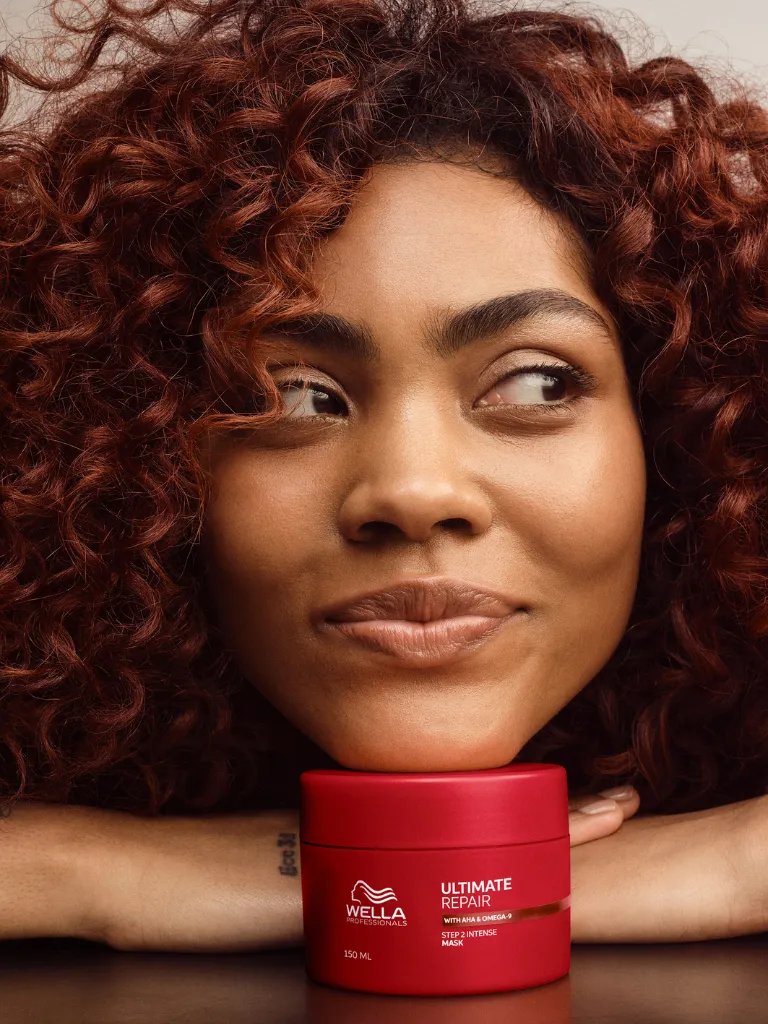 Model with dark brown, curly hair rests chin on tub of ULTIMATE REPAIR Mask.