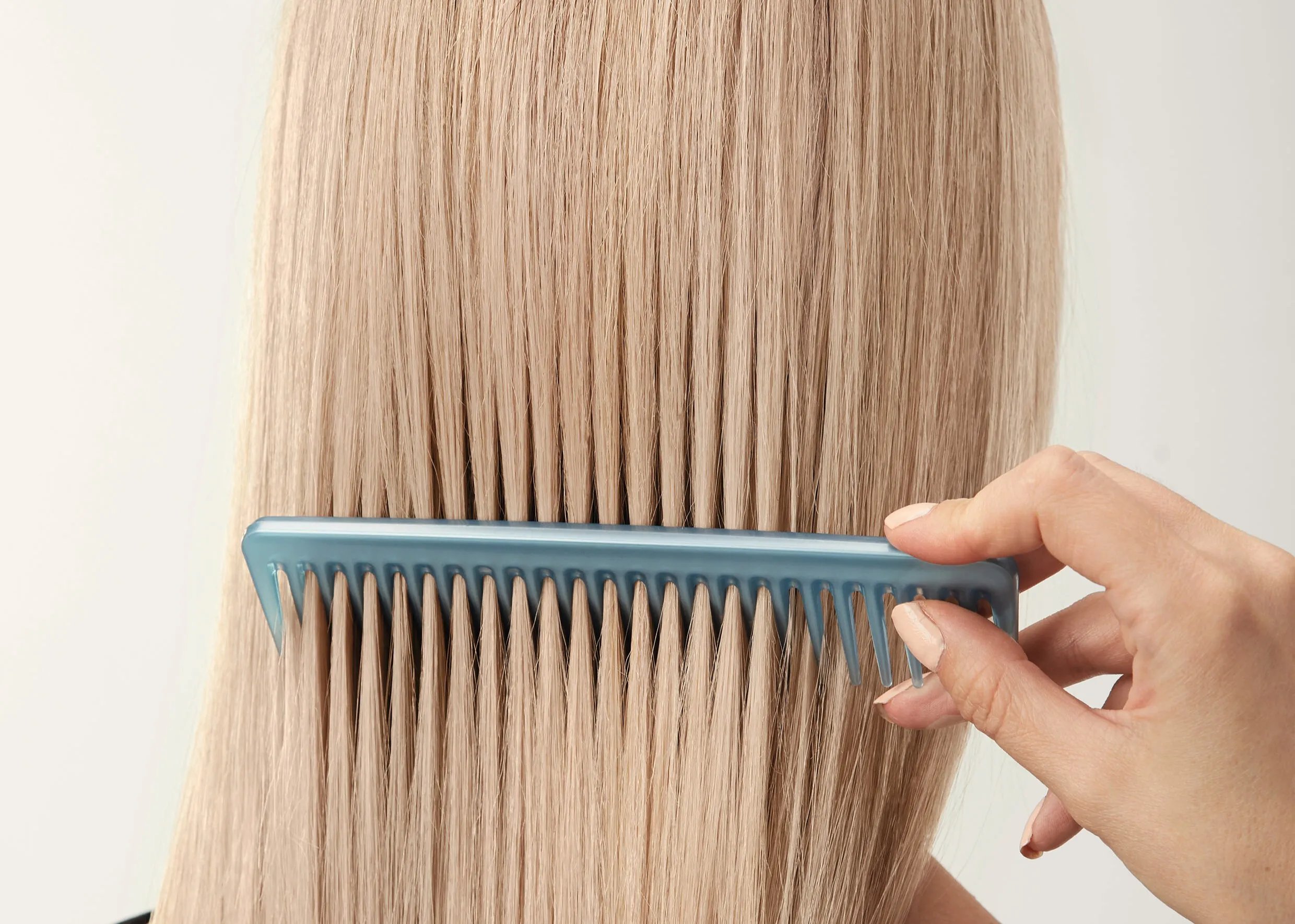 Close-up of long, straight, light-blonde hair being gently combed through with a blue comb, showing smooth strands against a neutral background