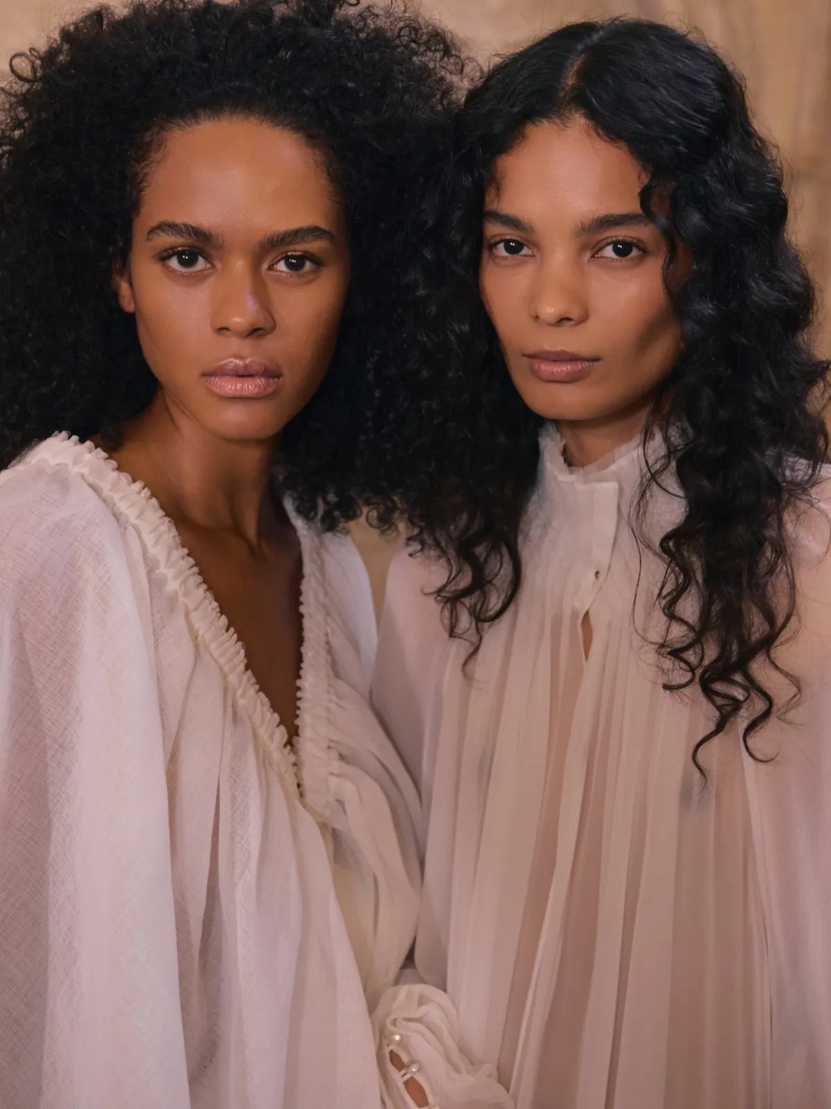 Two models backstage: one with full, voluminous, dark brown coils and one with dark brown, soft curls.