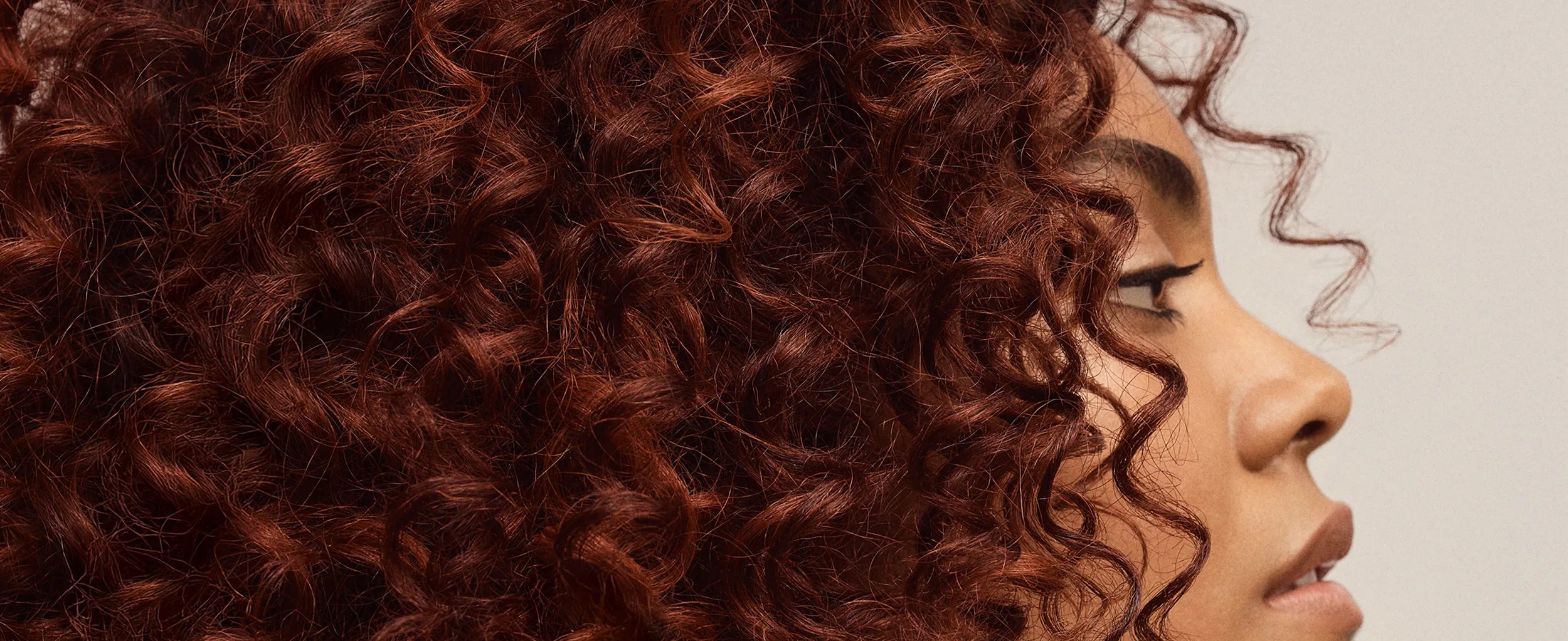 A profile view of a woman with tight, curly auburn hair against a neutral background.