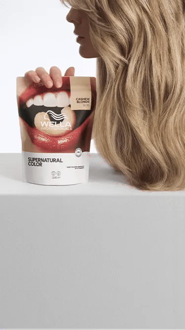 Εικόνα ενός μοντέλου σε προφίλ, που δείχνει τα ξανθά, κυματιστά μαλλιά του. Το μοντέλο κρατά στο χέρι του μια συσκευασία Wella Professionals Supernatural Color στην απόχρωση Cashew Blonde. Η εικόνα έχει τραβηχτεί σε λευκό φόντο.